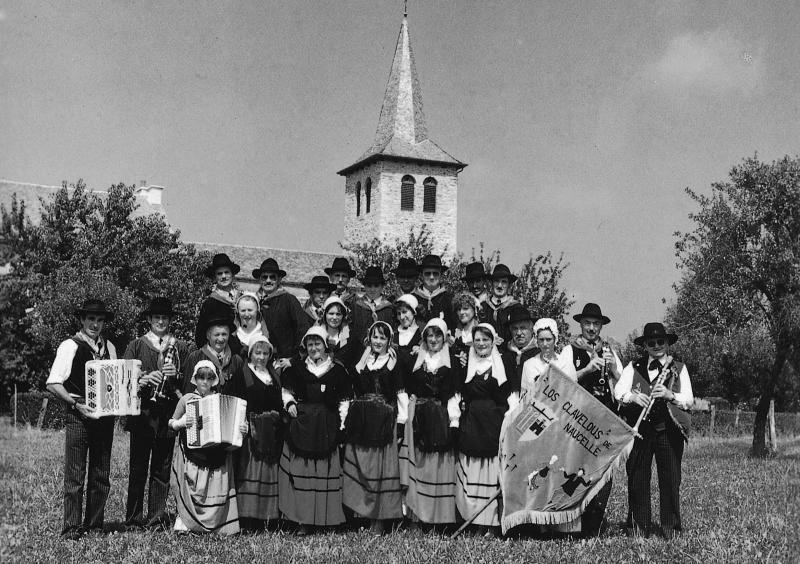 Groupe folklorique costumé, avec bannière avec inscription en occitan Los Clavelous [Clavelons] de Naucelle, dans une prairie (prada, prat) devant le clocher (cloquièr) de Cabanès